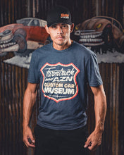 Person wearing a blue t-shirt with a museum logo in front of a backdrop with vintage cars.