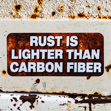 A rectangular metal sign with rounded corners featuring the text 'RUST IS LIGHTER THAN CARBON FIBER' in bold white letters. The sign has a distressed, rusted brown surface and is mounted on a similarly weathered, rust-speckled off-white metal background.