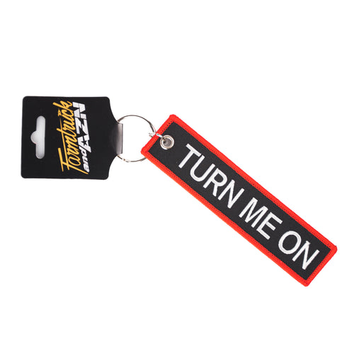 A keychain with a rectangular tag that has 'TURN ME ON' written on it, predominantly in white lettering with a black background, attached to a black and yellow card.