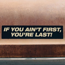 A bumper sticker with the phrase 'IF YOU AIN'T FIRST, YOU'RE LAST!' in black lettering on a brown background.