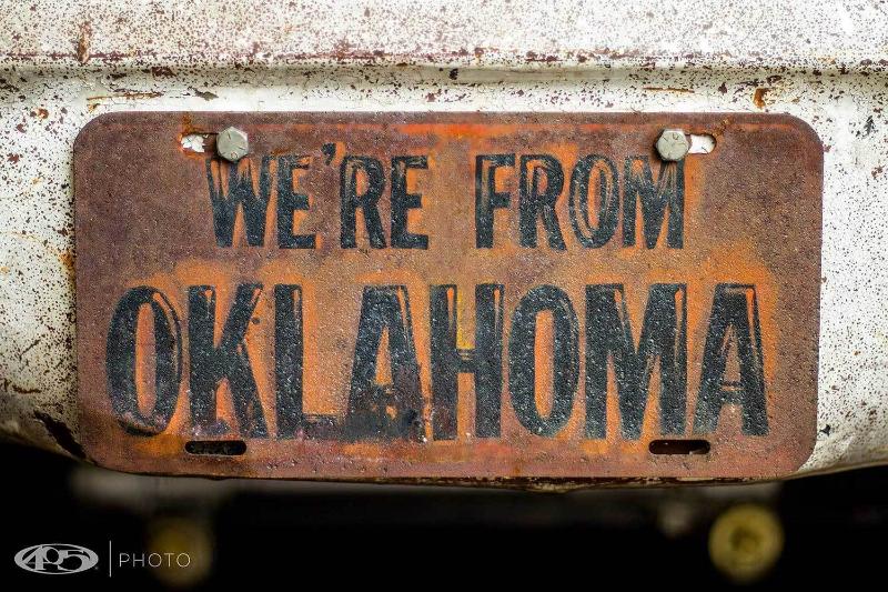 A custom full color printed 'We're From Oklahoma' vehicle license plate with rusty looking text and holes drilled for mounting on a vehicle.
